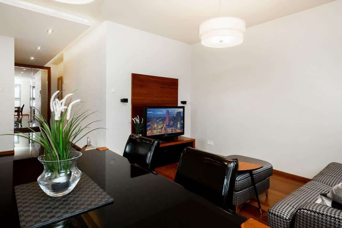 Ein modernes Wohnzimmer mit Esstisch, schwarzen Ledersesseln, einer Couch, einem Fernseher auf einem Holzständer und einer Vase mit weißen Blumen.