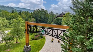 A wooden bridge with a green and white sign, surrounded by lush greenery and a building in the background.