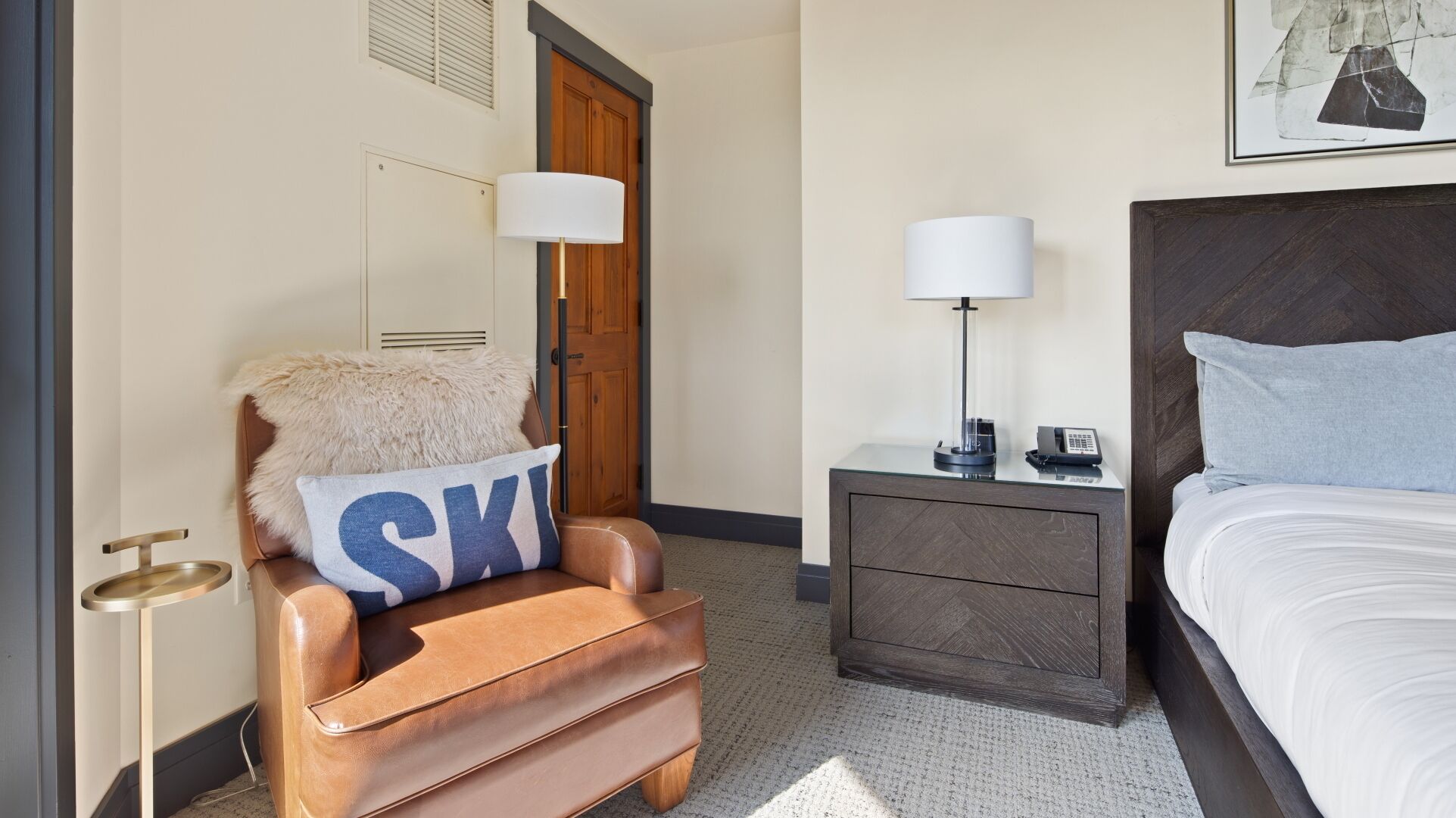 A hotel room with a brown leather armchair, a wooden door, a bedside table with a lamp, a bed, and a framed picture on the wall.