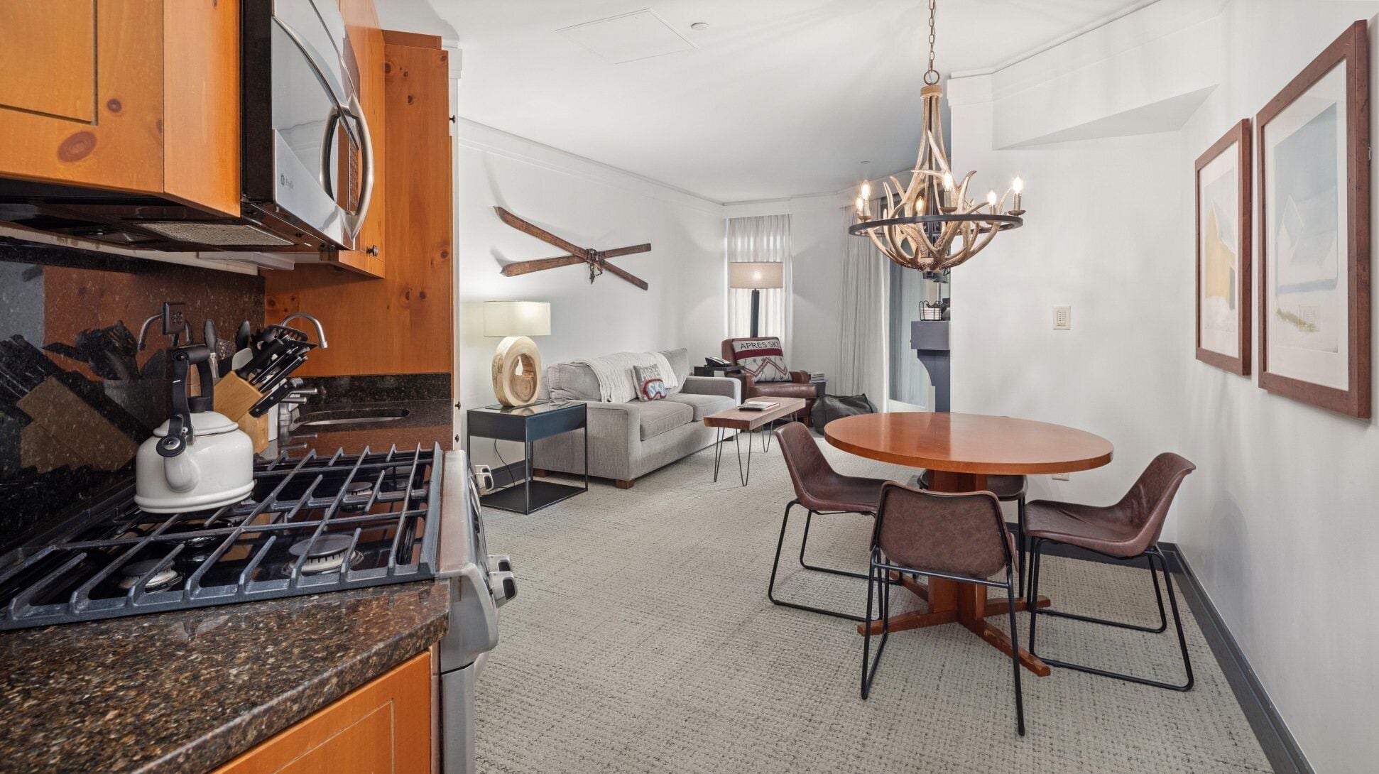 A modern kitchen with a gas stove, a kettle, and a coffee maker. There is a dining area with a round table and chairs. The living room has a sofa and a chair. A chandelier hangs from the ceiling.
