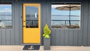 Un bâtiment avec une porte jaune, une fenêtre ornée d’un parapluie et un jardinier avec des plantes vertes.