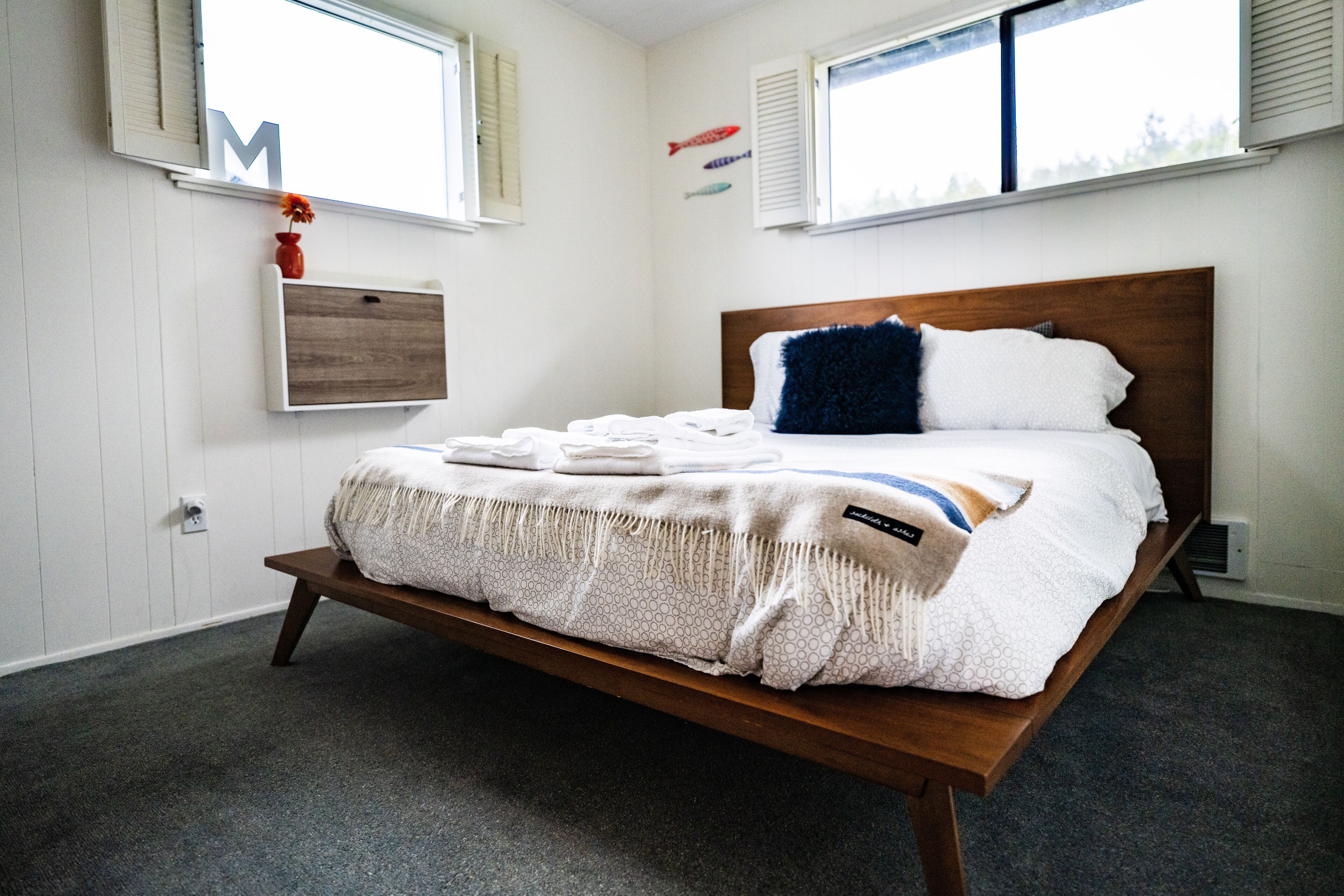 Een net opgemaakt bed met een houten hoofdbord, een klein plankje met een vaas, en een lichtschakelaar aan de muur.