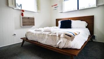 Een net opgemaakt bed met een houten hoofdbord, een klein plankje met een vaas, en een lichtschakelaar aan de muur.