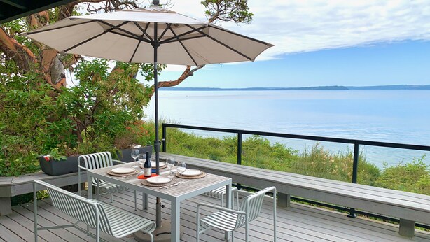 Un espace de restauration en plein air avec une table dressée pour quatre personnes, entourée de chaises blanches, sous un grand parasol.