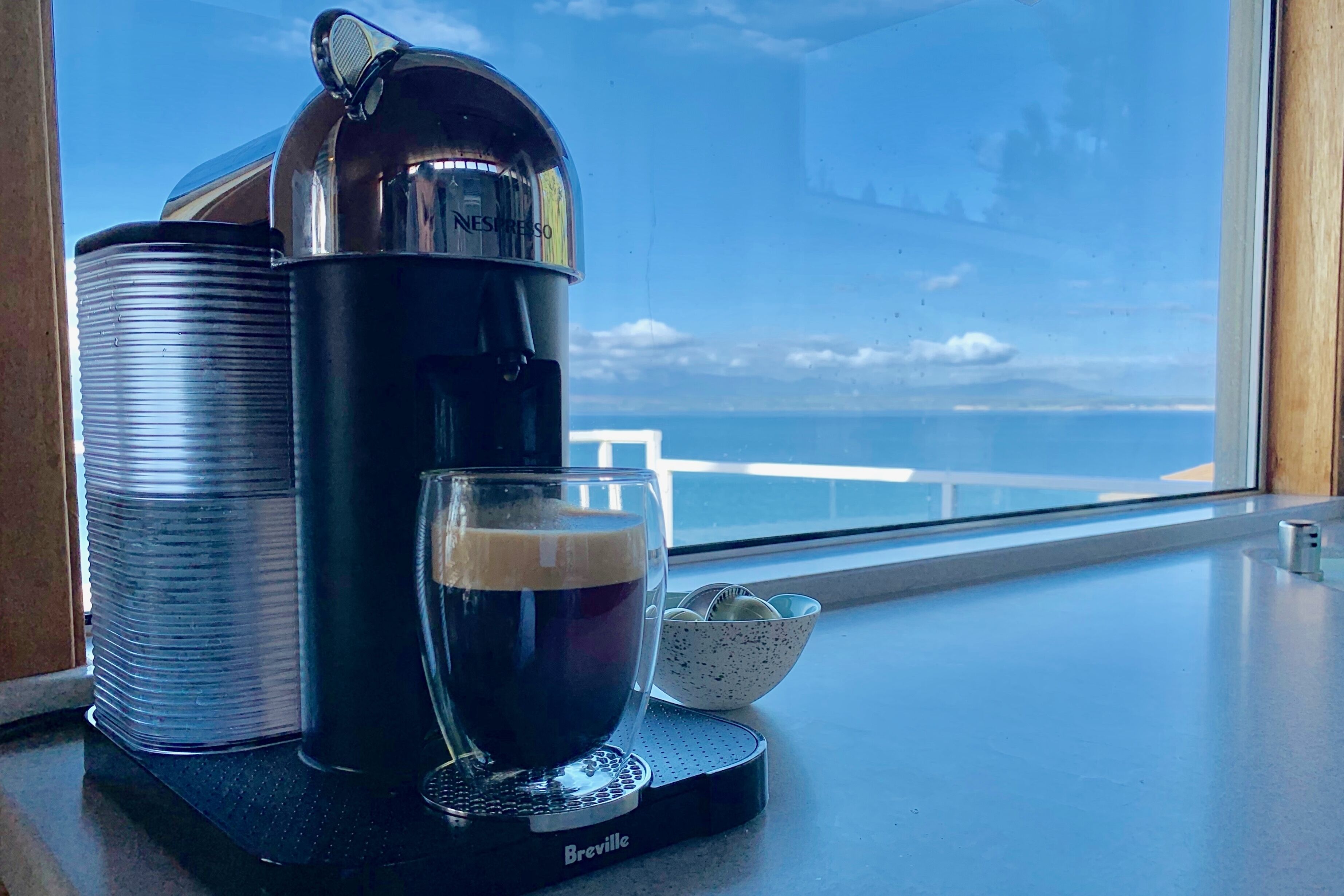 Une machine à café avec une tasse de café posée sur une table près d’une fenêtre donnant sur l’océan.