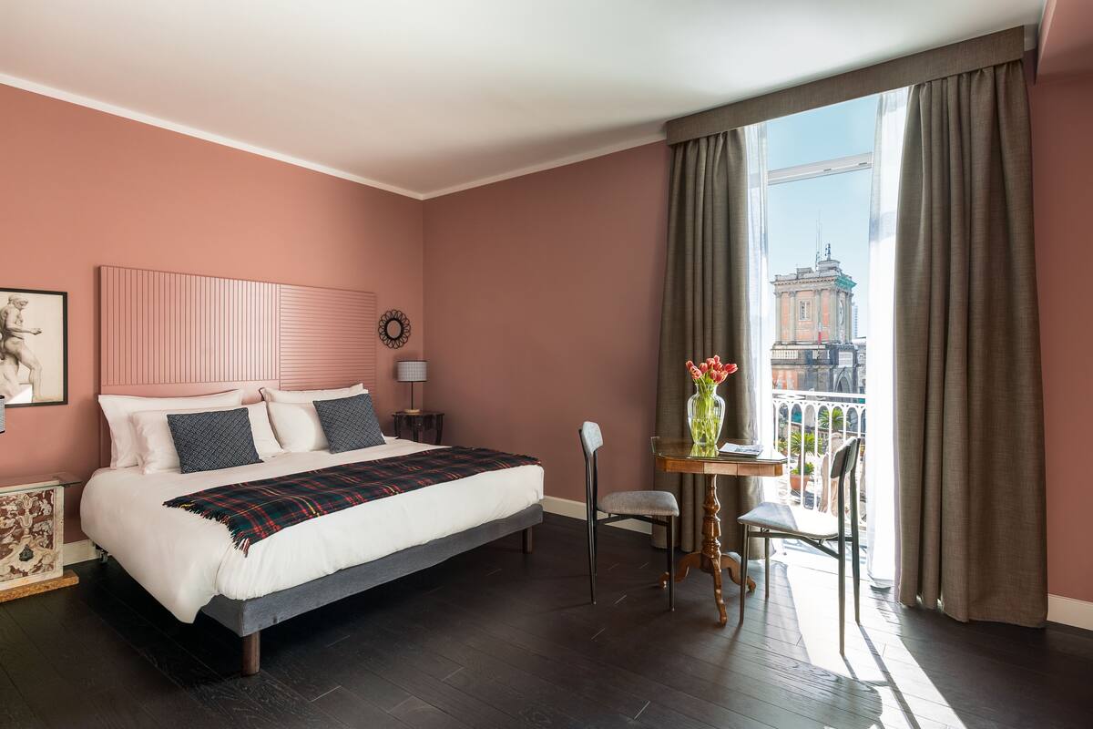 A bedroom with a large bed, a small table with a vase, and a view of a building through the window.