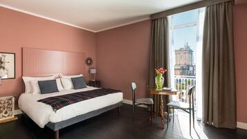 A bedroom with a large bed, a small table with a vase, and a view of a building through the window.