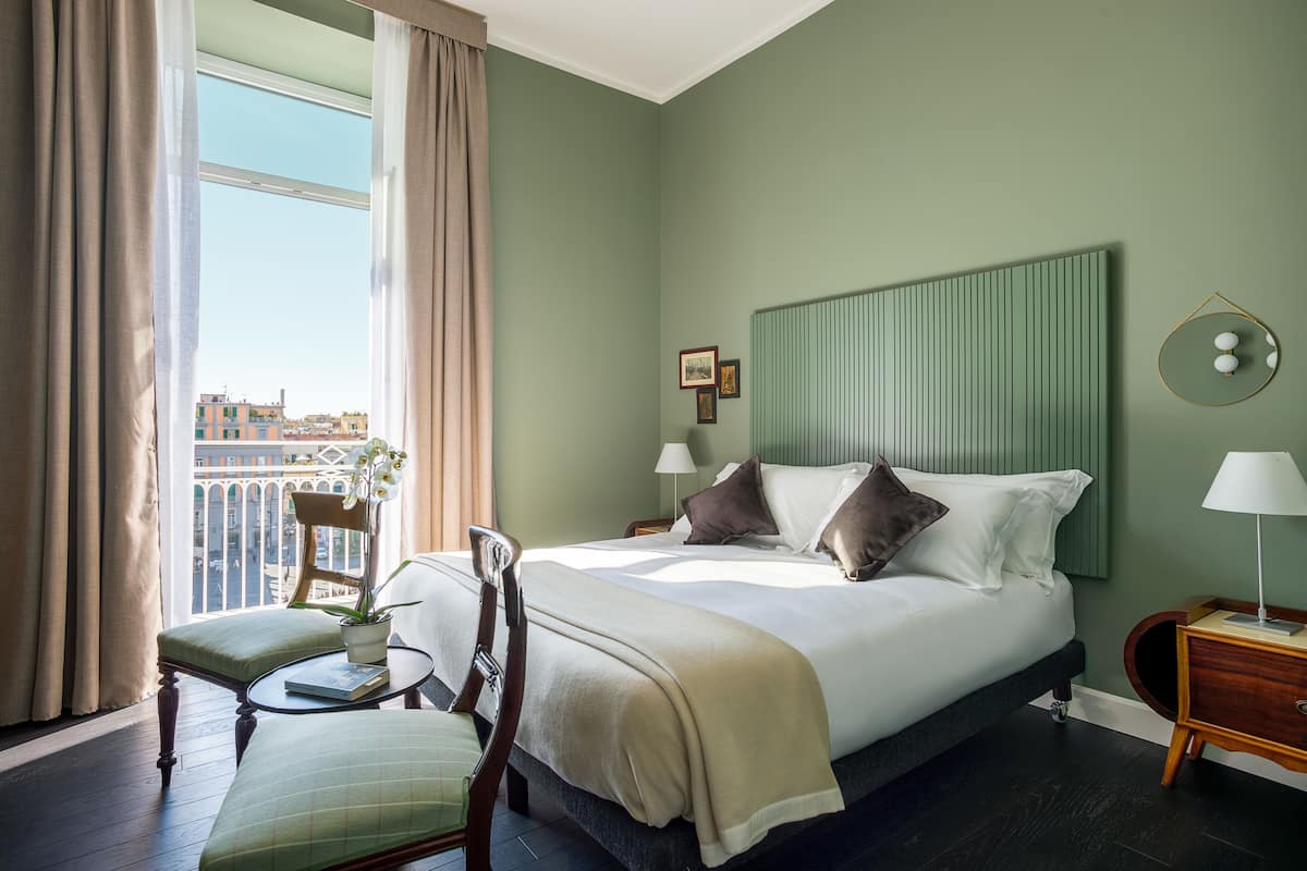 A hotel room with a large bed, a chair, a small table, a nightstand, and a view of buildings outside the window.