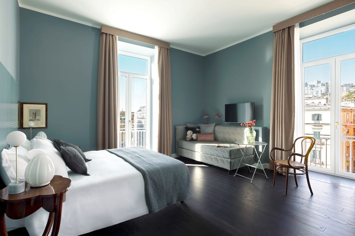 A hotel room with a large bed, a desk with a chair, a TV, and a view of the city through large windows.