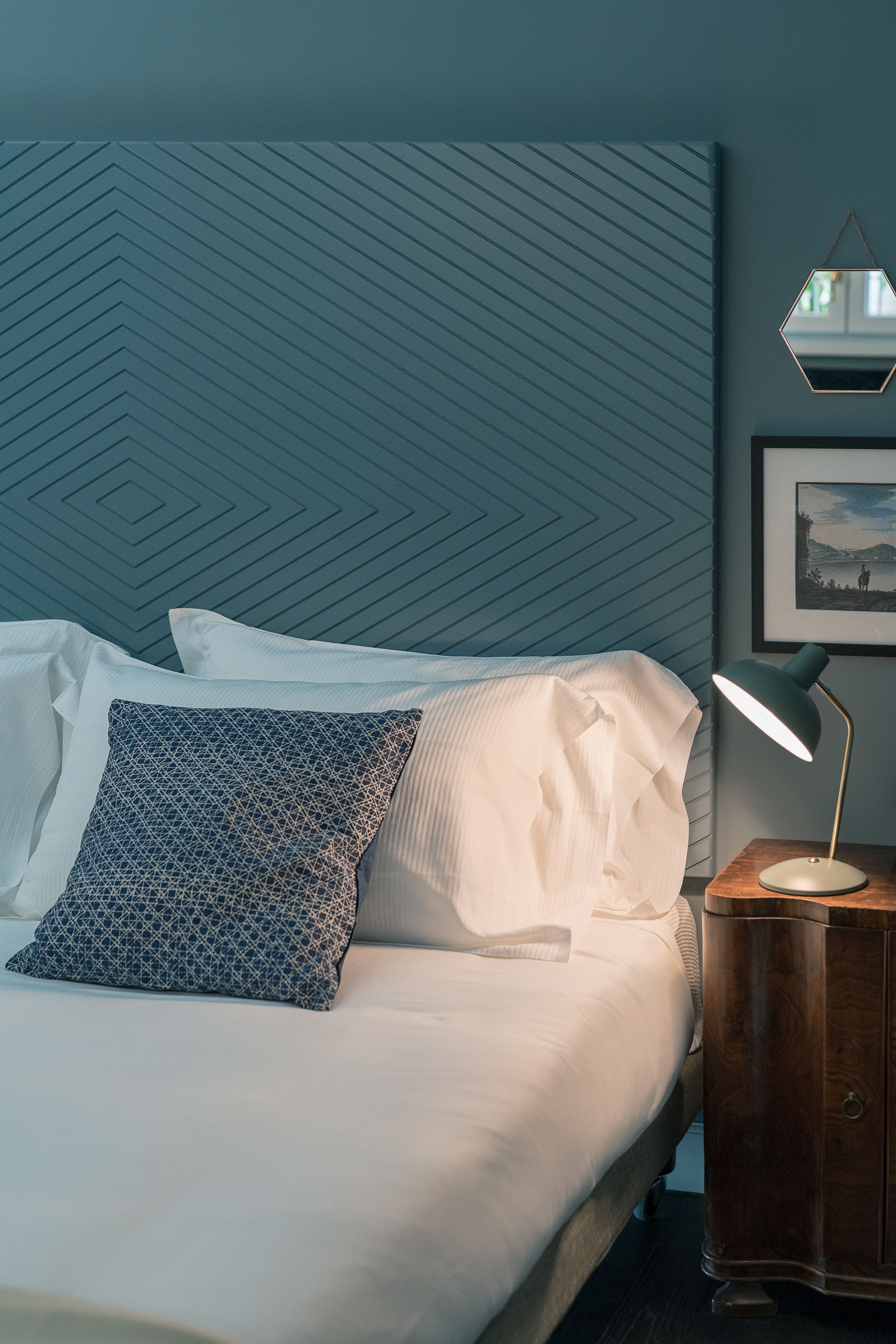 A neatly made bed with patterned pillows, a bedside table with a lamp, and a wall with a geometric pattern.