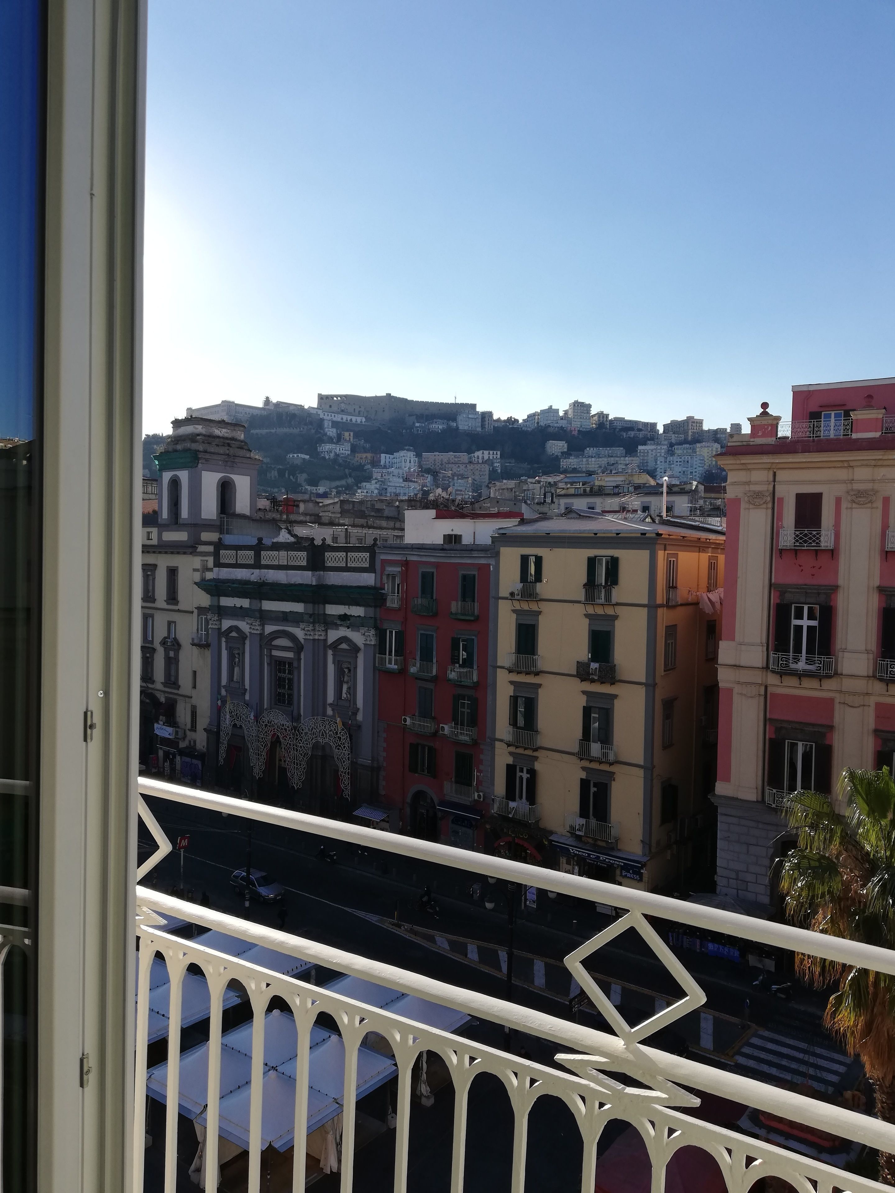 Un balcon offrant une vue sur des bâtiments aux couleurs vives et, au loin, une colline surmontée d’une structure ressemblant à un château.