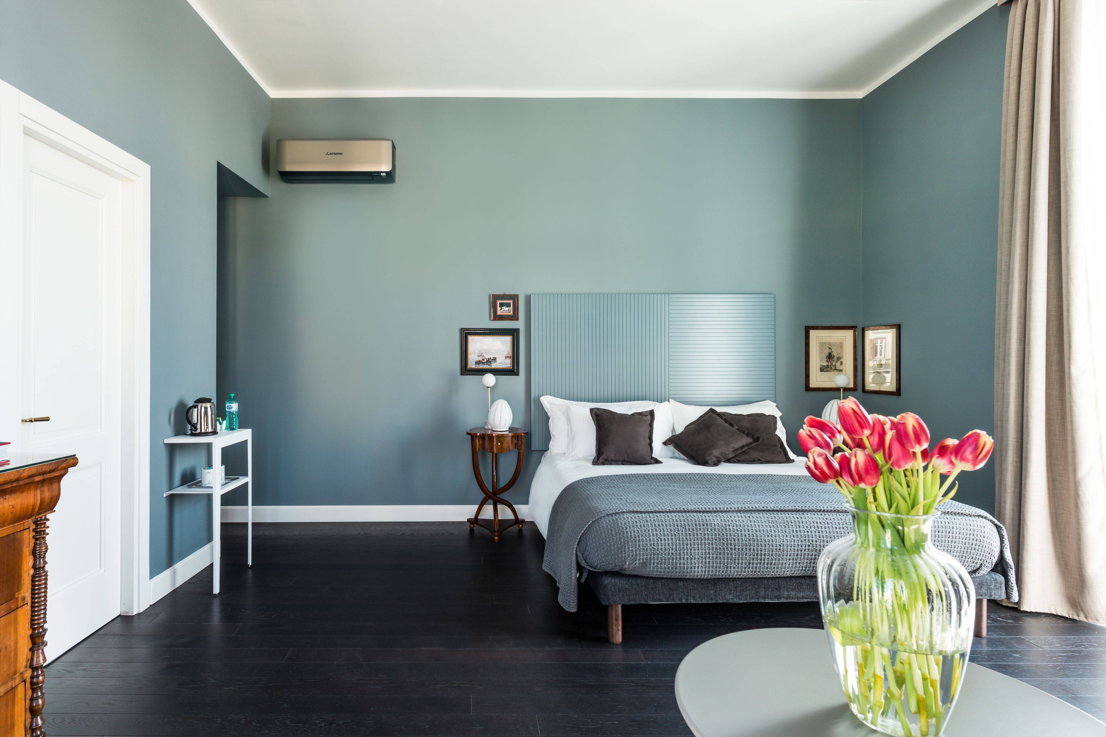 A bedroom with a bed, bedside tables, a nightstand, a vase with flowers, and a window with curtains.