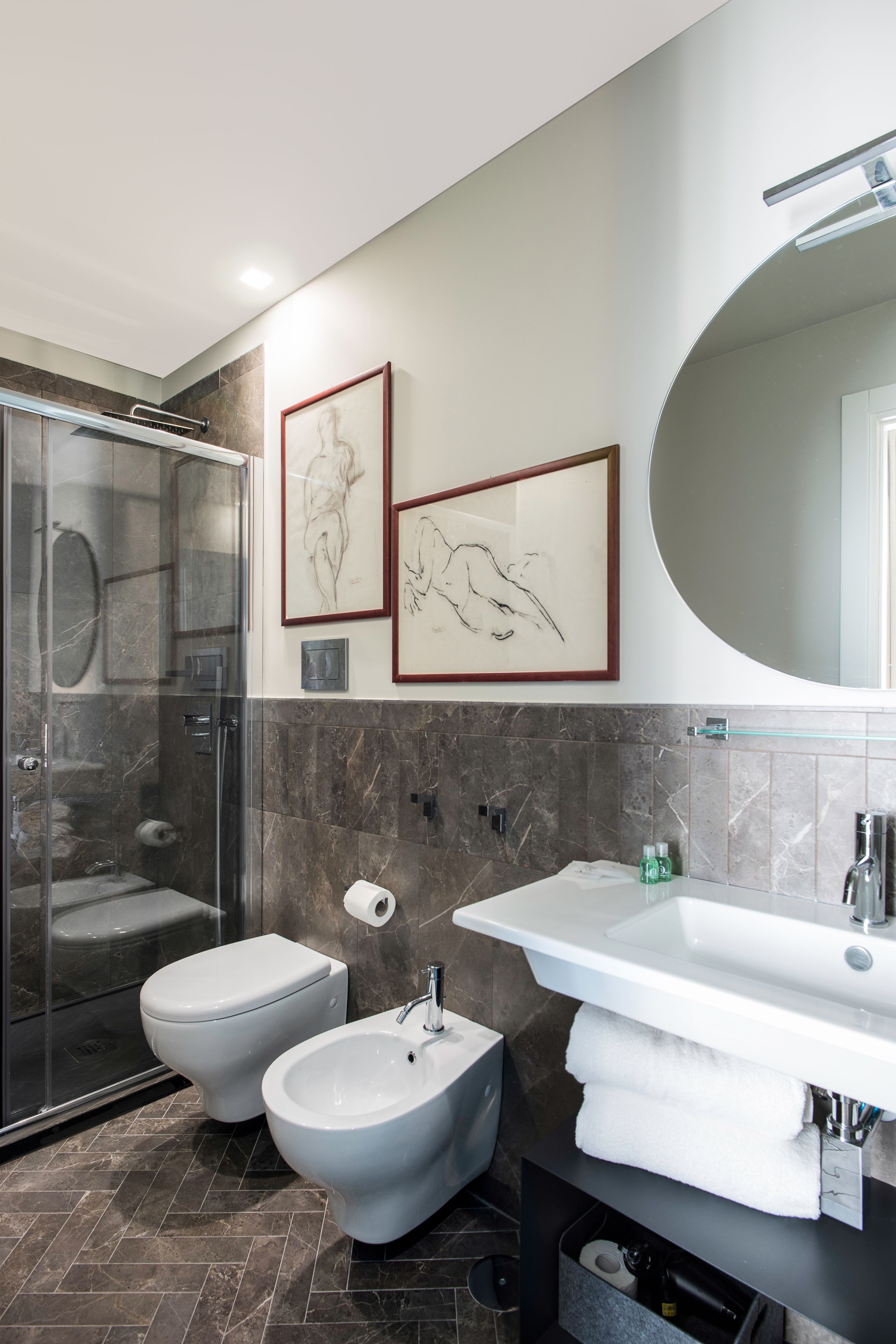 A modern bathroom with a glass-enclosed shower, a bidet, a large sink, and two framed artworks on the wall.