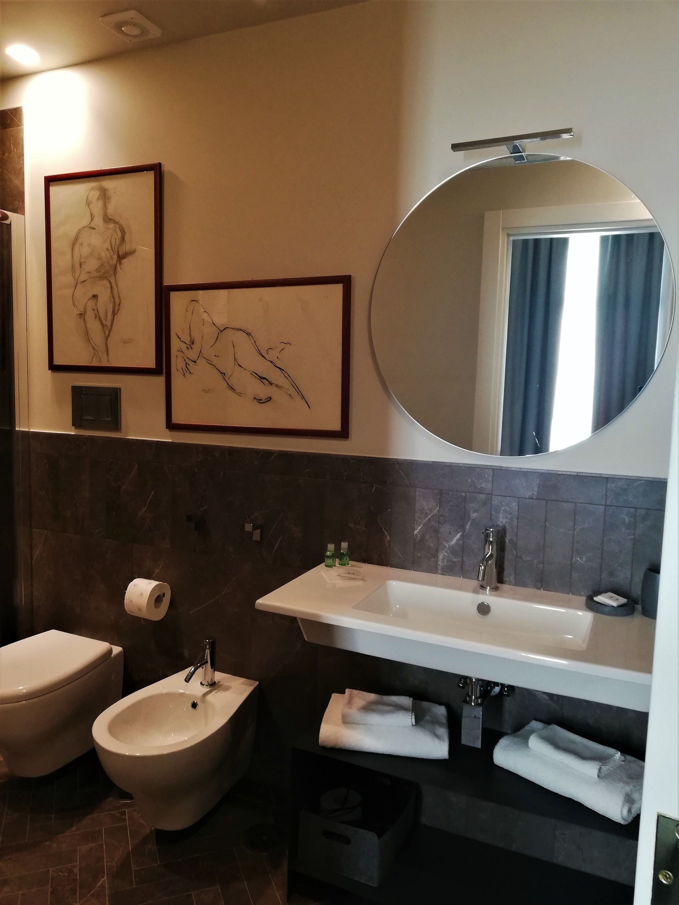 A modern bathroom with a bidet, toilet, and sink. There are framed artworks on the wall and a mirror above the sink.