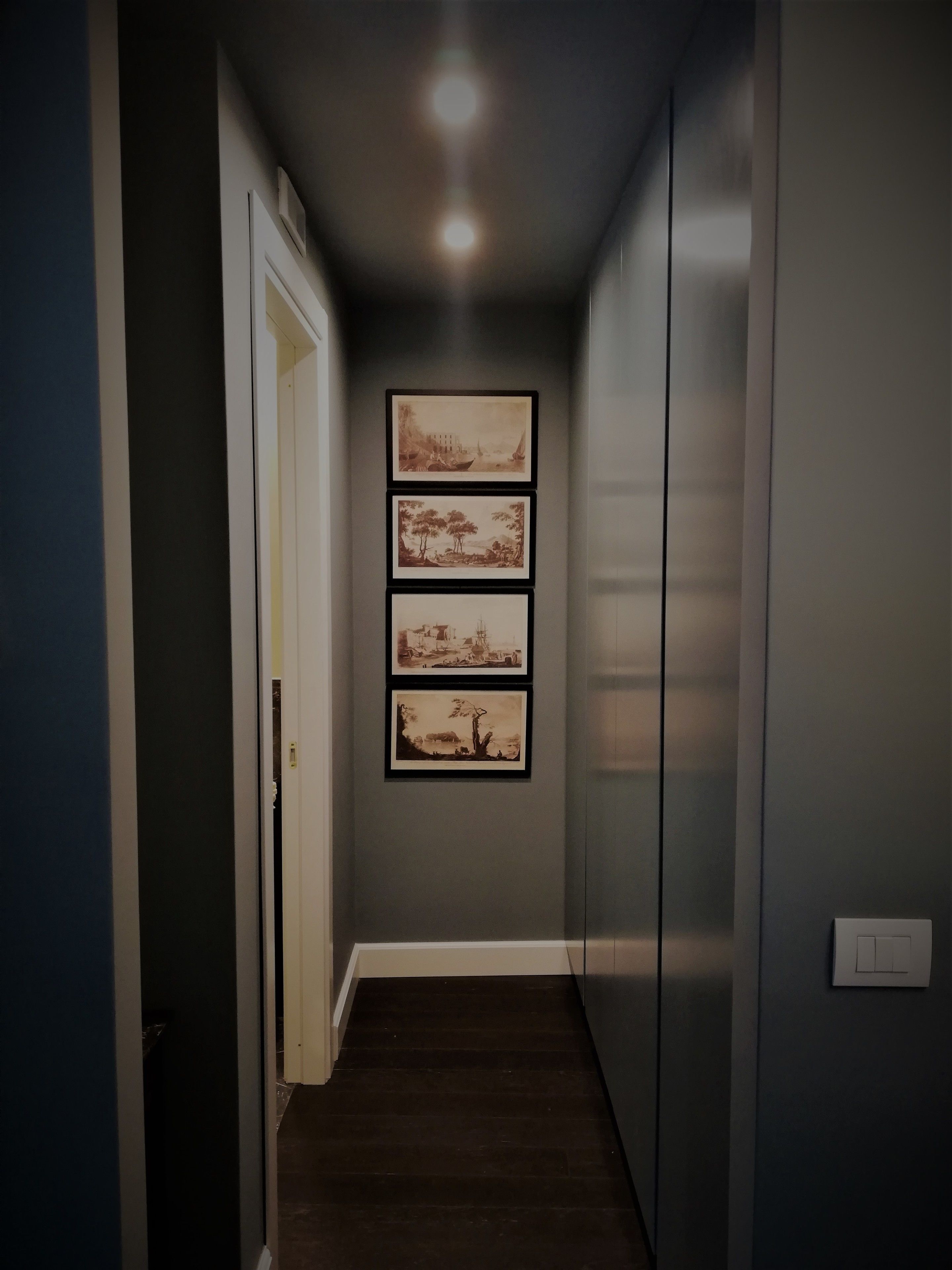 A narrow hallway with a dark wooden floor, light gray walls, and three framed pictures on the wall.