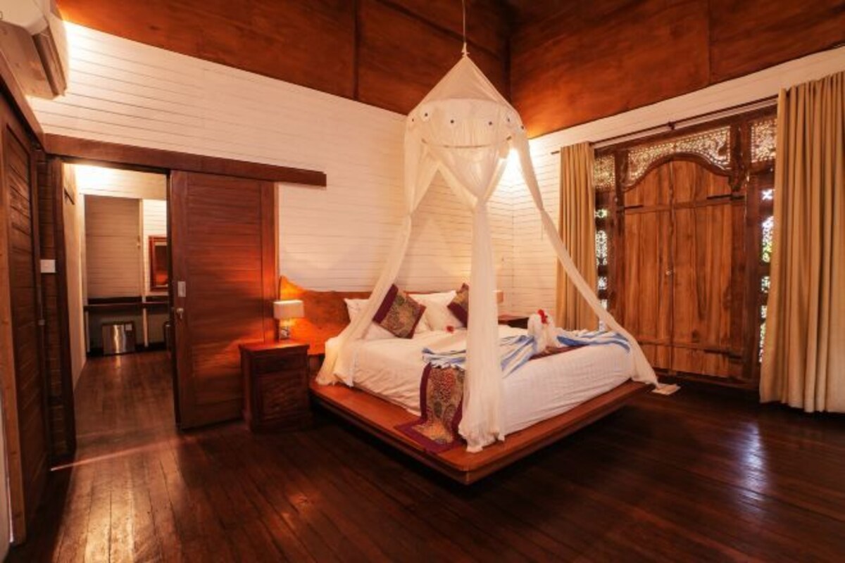 A bedroom with a canopy bed, wooden flooring, a wooden door, and a small bedside table with a lamp.