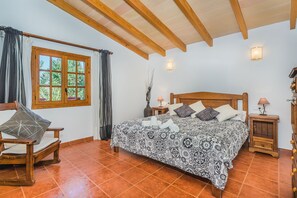 A bedroom with a large bed, wooden headboard, a window with curtains, a bedside table with a lamp, and a chair.