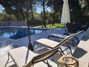 Zona de piscina con sillones de descanso y una pequeña mesa, rodeada por una valla y árboles.