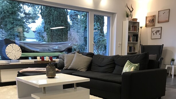 Ett modernt vardagsrum med en svart soffa, ett vitt soffbord och utsikt över träd genom stora fönster.