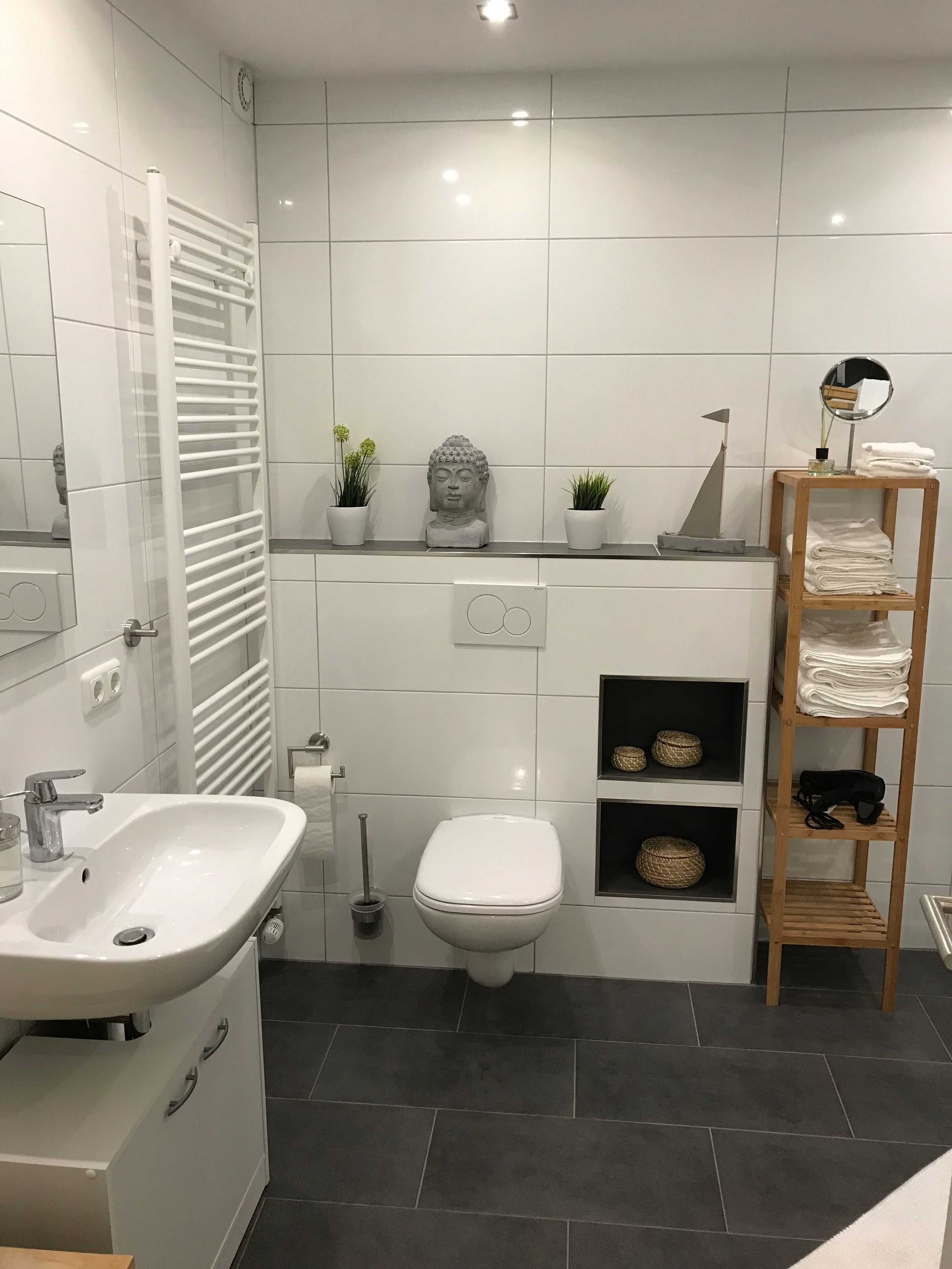 Un baño moderno con lavamanos, inodoro y toallero blancos. Hay dos plantas en macetas sobre el mueble y una pequeña estatua de Buda.