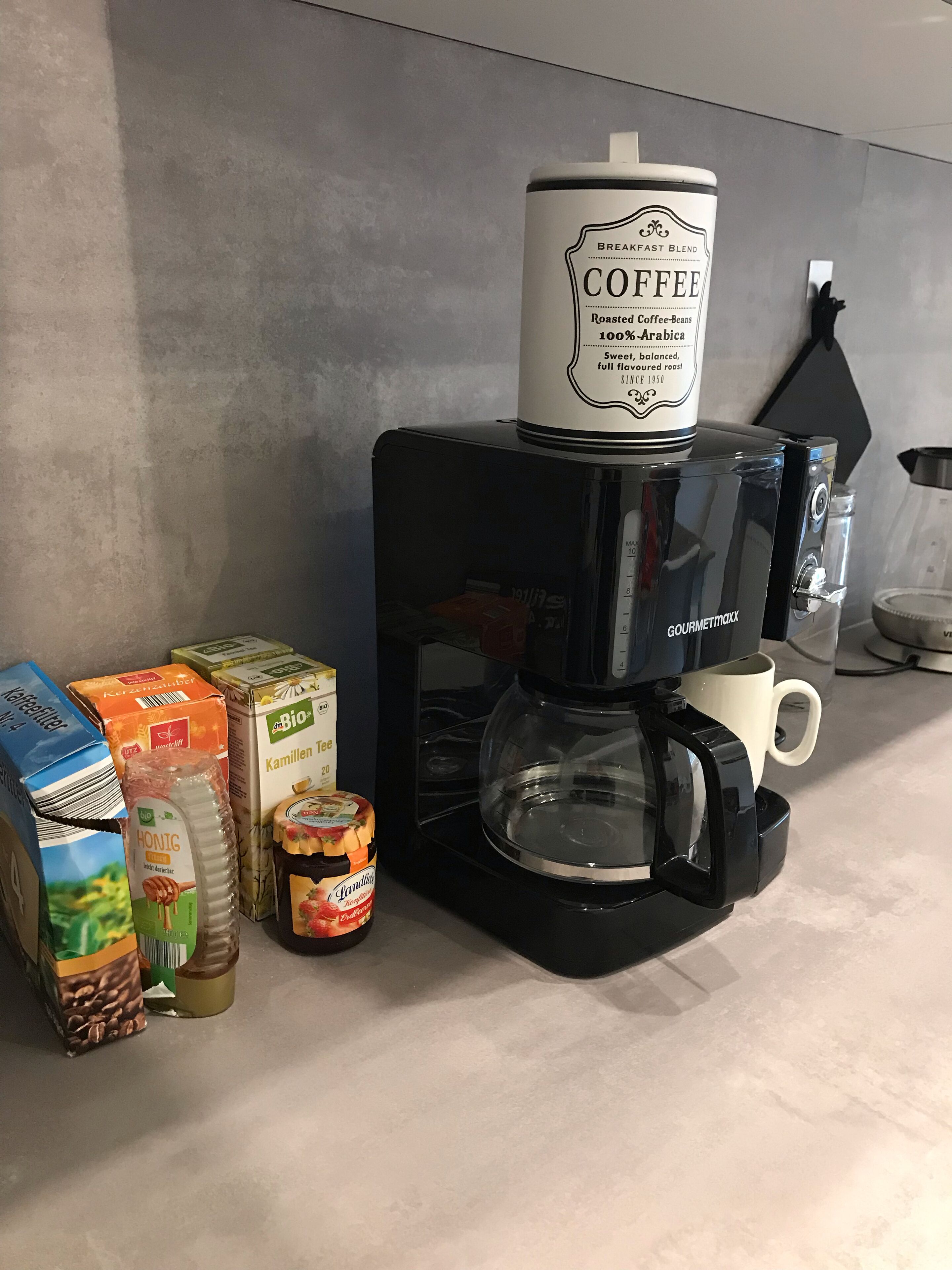 Une machine à café avec une verseuse sur le dessus, un mousseur à lait et divers produits à base de café et de thé disposés sur un plan de travail de cuisine.