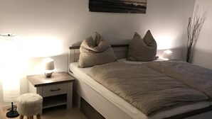 Ett sovrum med en säng, sängbord, en lampa, en liten pall och en tavla på väggen.