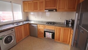 Une cuisine avec des meubles en bois, un réfrigérateur en acier inoxydable, un lave-vaisselle, une machine à laver, une cuisinière à gaz et une fenêtre avec des stores.