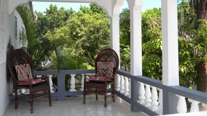 Un balcon abrité, agrémenté de deux fauteuils en osier, de balustrades blanches et d’une végétation luxuriante en arrière-plan.