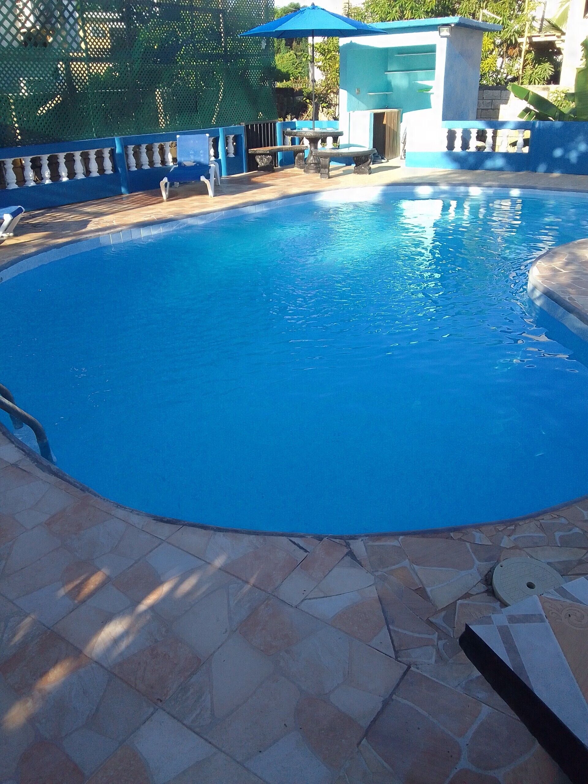 Une piscine avec une terrasse carrelée, un parasol bleu et une clôture blanche.