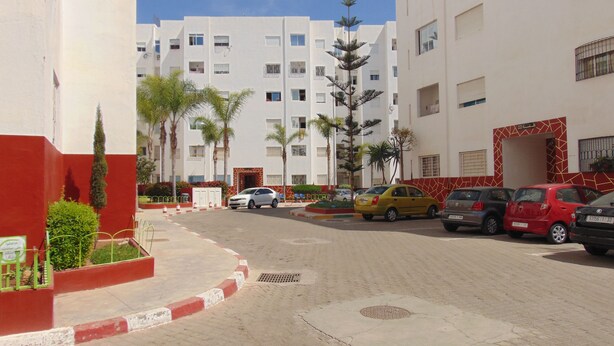 A street view with a row of parked cars, a building with a red and white facade, and a clear blue sky. - Suite Apartment Fawzi (Agadir)