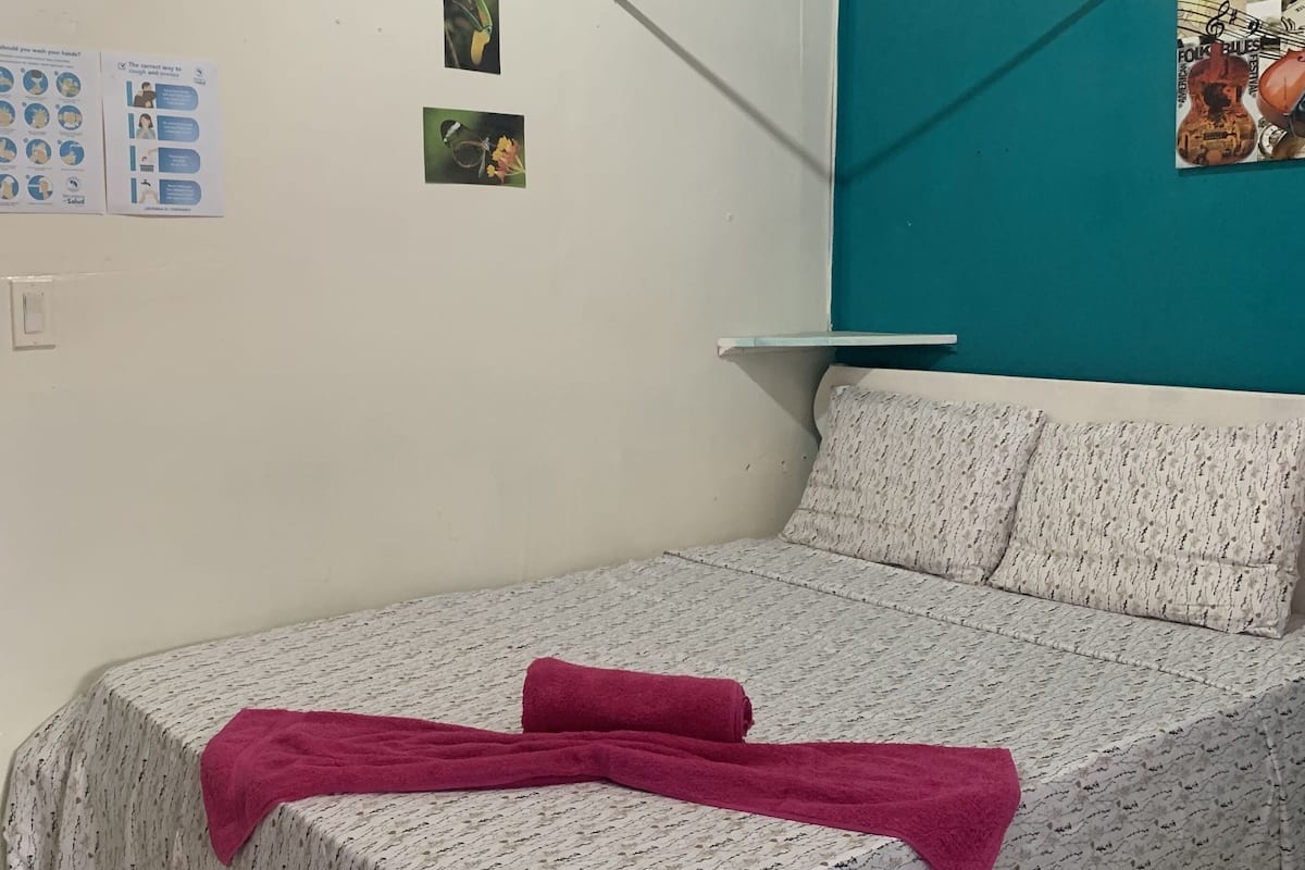 A single bed with a patterned bedspread and two pillows, a teal accent wall, a poster, and a bulletin board with various items.