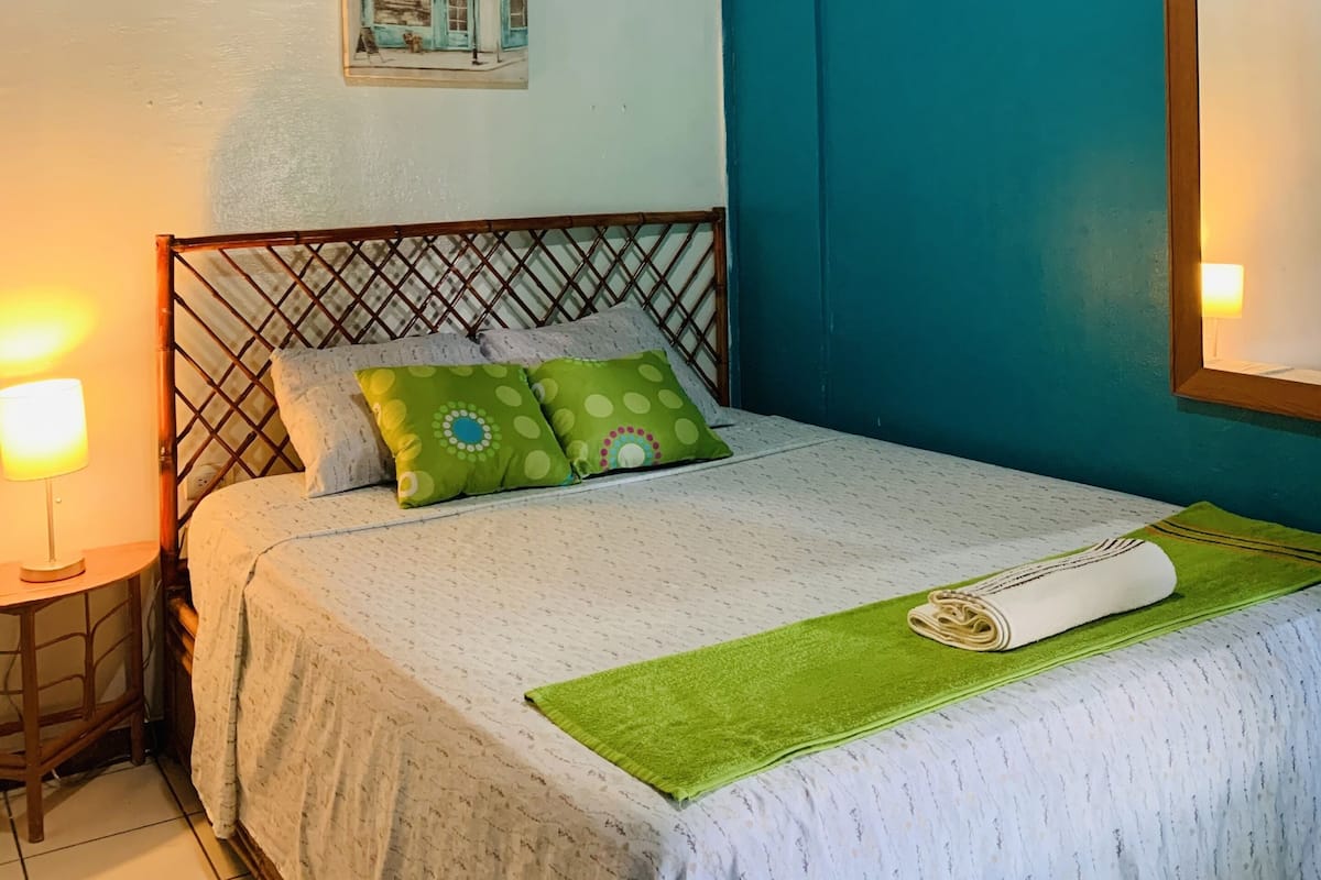 A neatly made bed with a green throw blanket and pillows, a wooden headboard, a wall-mounted air conditioner, and a framed picture of a street scene.