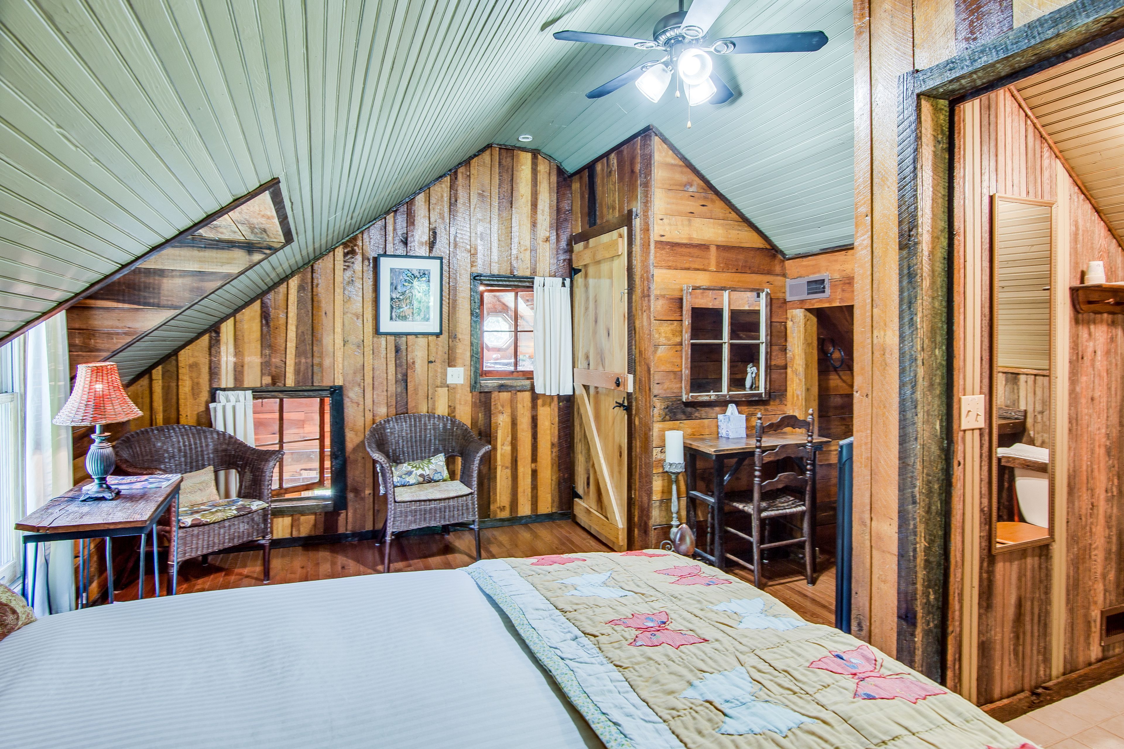 A cozy attic bedroom with wooden walls, a sloped ceiling, a bed with a floral quilt, a wicker chair, a small table with a lamp, and a ceiling fan.