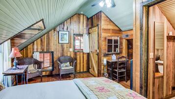 A cozy attic bedroom with wooden walls, a sloped ceiling, a bed with a floral quilt, a wicker chair, a small table with a lamp, and a ceiling fan.