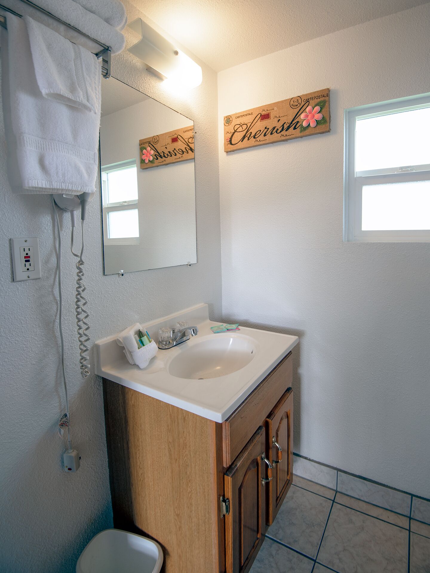 Un baño con lavamanos blanco, tocador de madera y un espejo. Se aprecia un perchero con una toalla blanca, un espejo pequeño y un cartel decorativo en la pared.