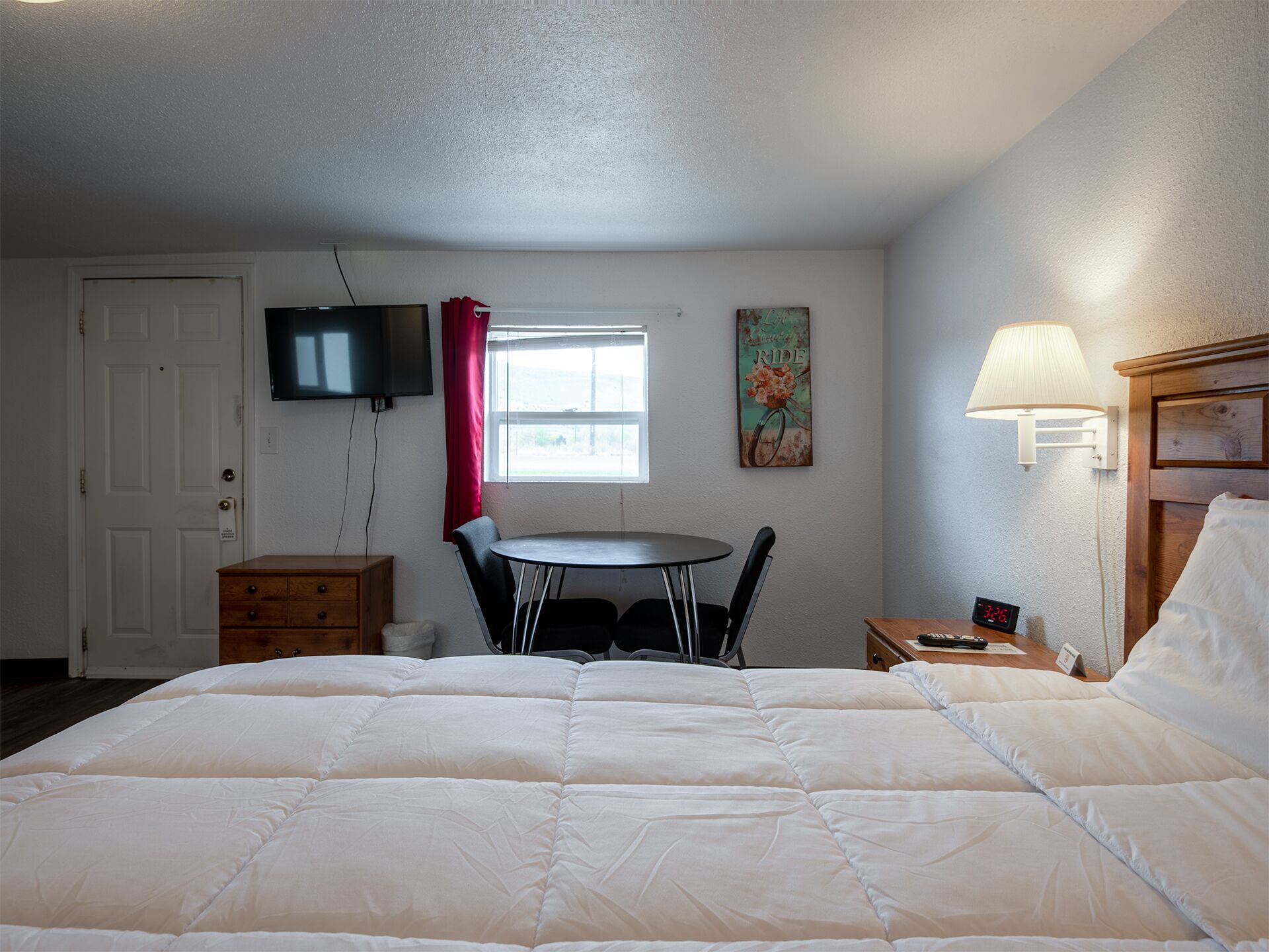 A hotel room with a bed, a small table with chairs, a TV, a window with curtains, a wall clock, and a lamp.
