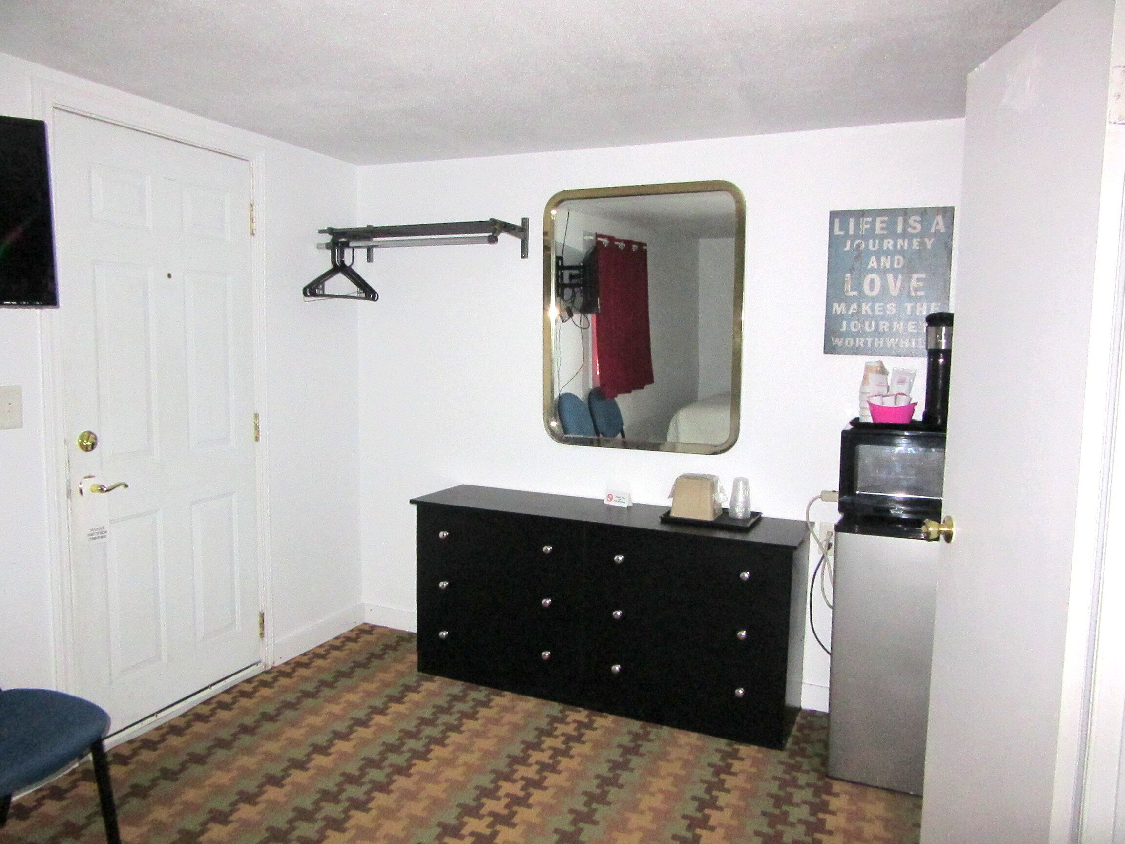 A small, compact room with a white door, a dresser with a mirror, a black heater, and a wall-mounted shelf with a kettle and cups.