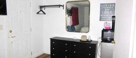 A small, compact room with a white door, a dresser with a mirror, a black heater, and a wall-mounted shelf with a kettle and cups.