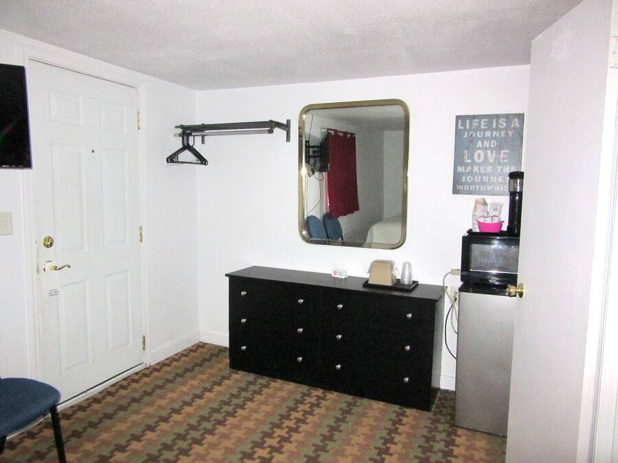 A small, compact room with a white door, a dresser with a mirror, a black heater, and a wall-mounted shelf with a kettle and cups.