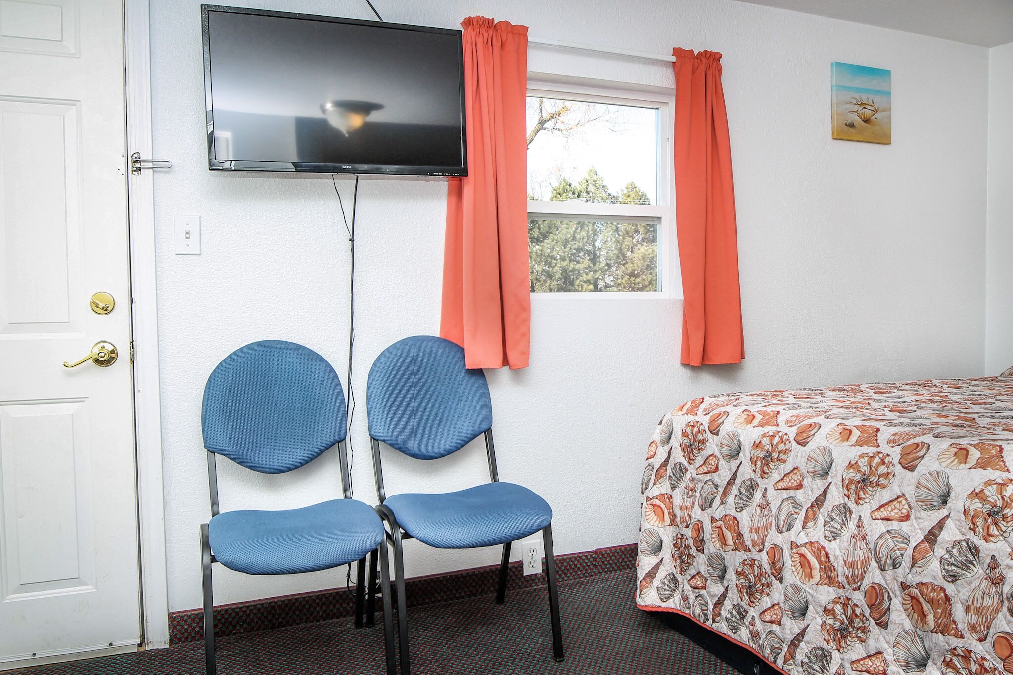 A room with a bed, two blue chairs, a television, and a window with orange curtains.