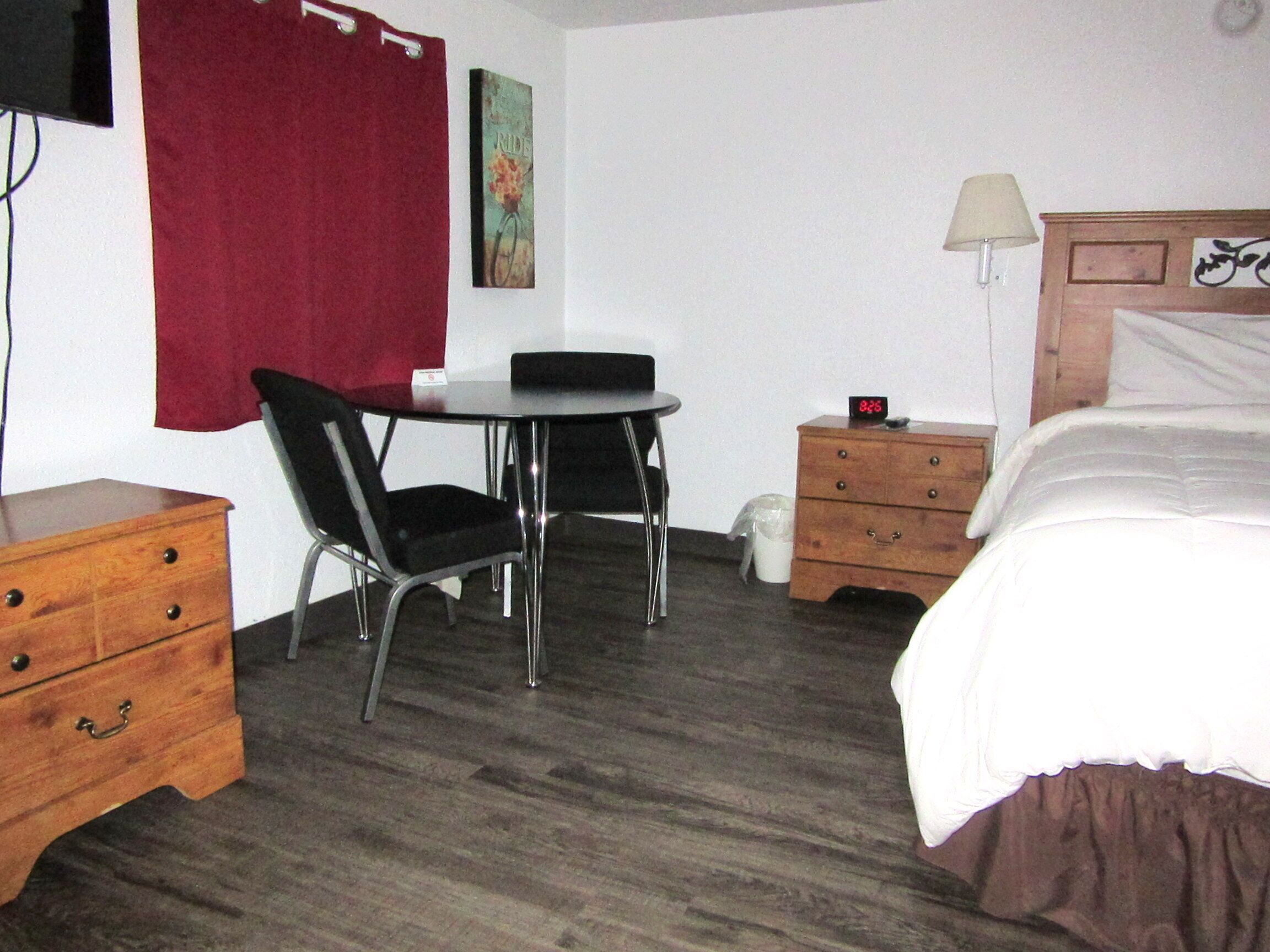 Une chambre d’hôtel avec une commode en bois, une chaise noire, une petite table ronde, un lit avec une literie blanche, une table de chevet avec une lampe et un tableau accroché au mur.