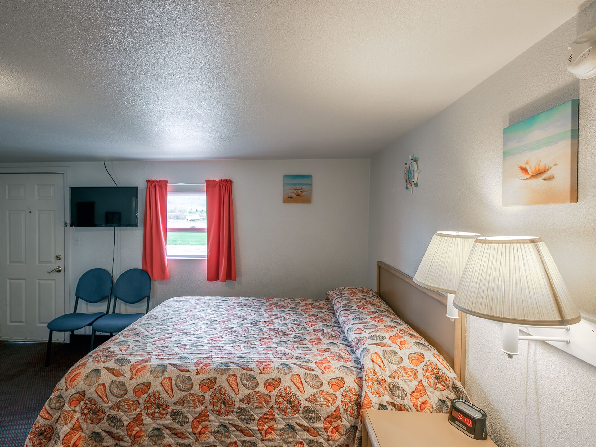Une chambre d’hôtel comprenant un lit, deux chaises, un bureau, une lampe, un téléviseur et une fenêtre avec des rideaux.