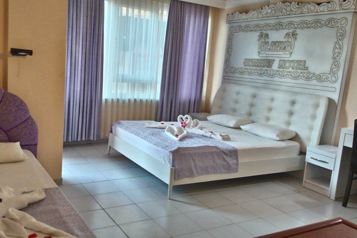 A hotel room with a bed, a nightstand, a headboard with a logo, and a window with curtains.
