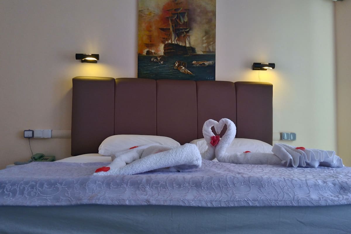 A hotel room with a large bed, two wall-mounted lights, a painting of a ship, and a red flower on the pillow.