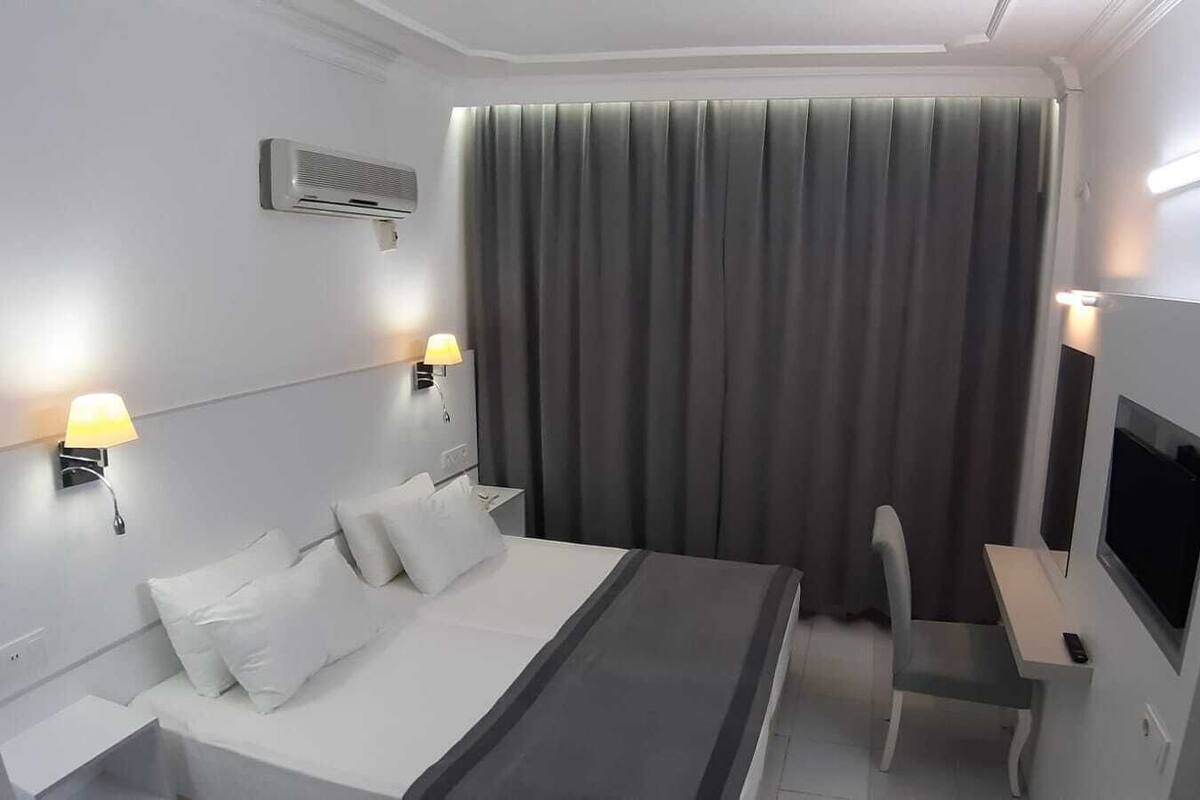 A hotel room with a bed, two bedside tables with lamps, a TV, and a large window with curtains.