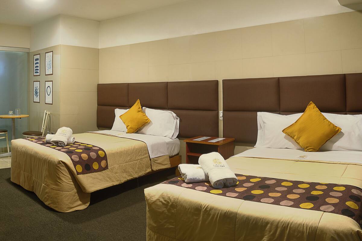A hotel room with two beds, each with a brown headboard, white linens, and a yellow pillow. There are towels on the beds and a small wooden nightstand between them.