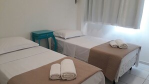 A hotel room with two single beds, each with a brown bedspread and white pillows. There is a teal side table between the beds, and a window with sheer curtains. - Ed. Bosque das Azaleias (Salvador)
