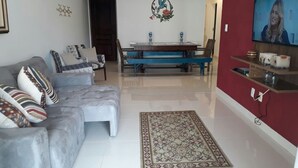 A living room with a grey sofa, a patterned rug, a dining table with chairs, and a TV on a wooden shelf. - Ed. Bosque das Azaleias (Salvador)