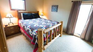 Un dormitorio con estructura de cama de madera, colcha, mesita de noche con lámpara, ventana con cortinas y un letrero en la pared.