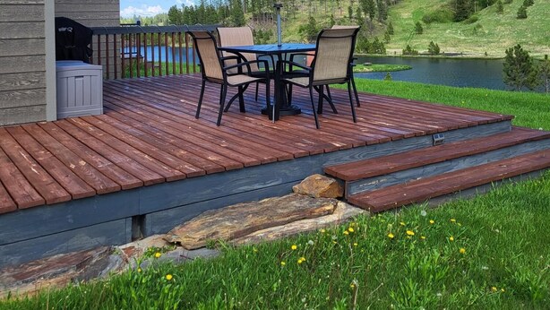 Una terraza de madera con una mesa y sillas, con vista a un lago y colinas boscosas.